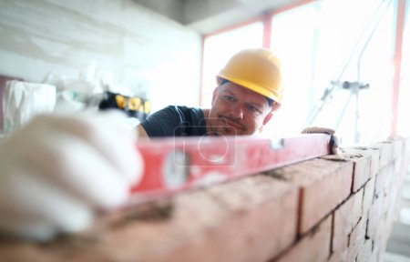 Foto de Retrato de constructor barbudo usando guantes duros para mantener las manos ilesas. Hombre pedante utilizando una herramienta especial para medir la altura y el nivel de la pared de ladrillo. Concepto de construcción - Imagen libre de derechos