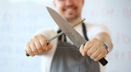 Photo for Professional Kitchen Knife Sharpening Photography. Chef Whetting Steel Blade Manually on White Background. Standing Man Holding in Hands Sharp Kitchenware for Cooking Partial View Horizontal Shot - Royalty Free Image