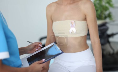 Téléchargez les photos : Le médecin mammologue tient une radio dans le presse-papiers près du patient avec un torse bandé dans le gros plan de la clinique. Lutte contre le cancer du sein concept. - en image libre de droit