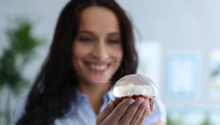 Téléchargez les photos : Gros plan d'une jeune femme souriante heureuse tenant un implant en gel. Travailleur médical joyeux à l'air surpris. Rendez-vous clinique. Médecine moderne et concept de correction corporelle - en image libre de droit