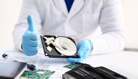 Photo for Male repairman wearing blue gloves is holding hard drive from computer laptop in hands show OK thumbs up. Performs diagnostics performs urgent repairs recovery of lost data during deletion HDD closeup - Royalty Free Image