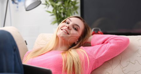 Téléchargez les photos : Jeune belle femme blonde allongée sur le canapé dans le salon tenir tablette dans les bras concept matin ensoleillé - en image libre de droit