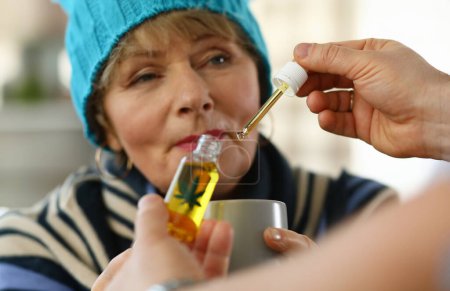 Téléchargez les photos : Malade femme âgée à la maison prend gouttes tasse de cannabis. Effets secondaires drogues. Médecine froide pour personnes âgées. Faible système immunitaire dans la vieillesse. Utilisez le médicament à des doses thérapeutiques. Danger pour les personnes âgées - en image libre de droit