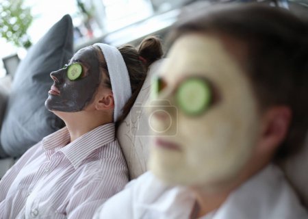 Téléchargez les photos : Les gens sont allongés sur le canapé avec des visages de masque cosmétique. Homme et femme couchent avec un masque sur les concombres visages. Week-end détente. Masques de tapis pour la peau. Concombres sur les yeux pour hydrater les paupières - en image libre de droit