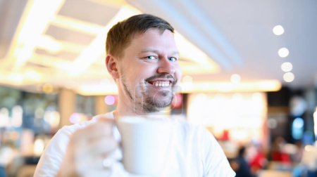 Téléchargez les photos : Homme souriant assis dans un café avec une grande tasse de café. L'homme se réjouit d'ouvrir des restaurants et des zones d'été après une pandémie. Attitudes positives. Ascenseur émotionnel d'une promenade du soir - en image libre de droit