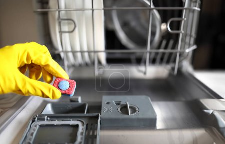 Foto de Guante manos sosteniendo la tableta lavavajillas lavado. Establecer el modo de lavado requerido. Los productos contienen componentes de espuma mínimos. Detergente concentrado. Secado efectivo. Limpiamos todos los platos de superficies - Imagen libre de derechos