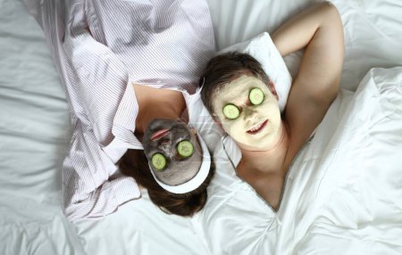 Téléchargez les photos : Couple couché sur le lit avec masque cosmétique sur le visage. Mari et femme s'amusent à la maison pendant la quarantaine. Les gens mettent des concombres sur leurs yeux pour hydrater leurs paupières. Détente pendant l'auto-isolement - en image libre de droit