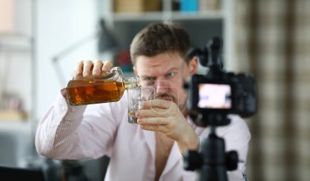 Téléchargez les photos : Caméra frontale Shabby Man et se déverse du whisky. Rowdy favorise l'ivresse. La personne est accro à l'alcool. Le gars s'inquiète pour le coronavirus et boit de l'alcool devant la caméra. - en image libre de droit