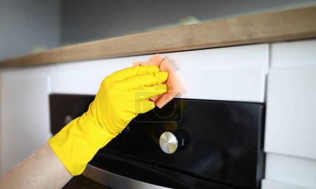 Close-up of persons hand in yellow protective gloves cleaning kitchen appliance with pink rag. Modern cookstove with display. Handyman and cleaner service concept