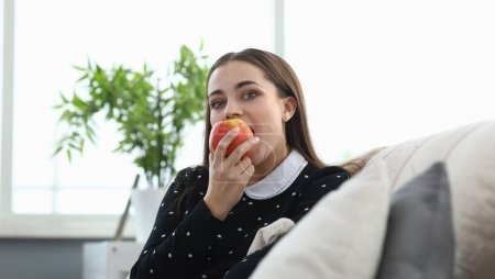Foto de Retrato de atractiva hembra mordiendo jugosa fruta de manzana. Acogedor interior del hogar. Mujer joven y bonita comiendo vitamina sentada en el sofá. Concepto de ocio y alimentación saludable - Imagen libre de derechos