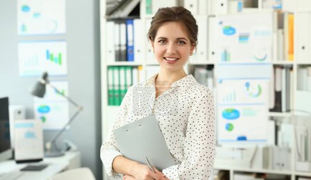 Beautiful smiling woman standing in office holding document clipped to pad looking in camera headshot