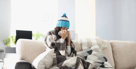 Kranke Frau mit warmem Hut bläst zu Hause ihre Nase in Papierserviette. Behandlung saisonaler Erkältungen zu Hause