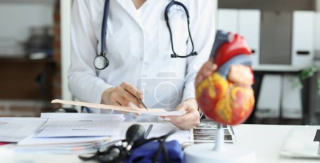 Doctor cardiologist examining cardiogram against background of artificial heart model closeup. Diagnosis of myocardial infarction and angina pectoris concept