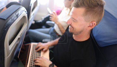 Junger Mann fliegt im Flugzeug und tippt auf Laptop-Tastatur. Fernkommunikationskonzept