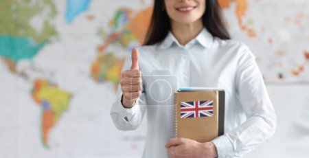 Frau hält englische Lehrbücher auf dem Hintergrund der Weltkarte und zeigt Daumen hoch. Konzept für Englischunterricht