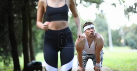 Photo for Tired guy does not have time to run with girl. Morning jogging helps body wake up and start vital mechanisms in it. Man leaned over and restores breath. Running in morning will help lose weight. - Royalty Free Image
