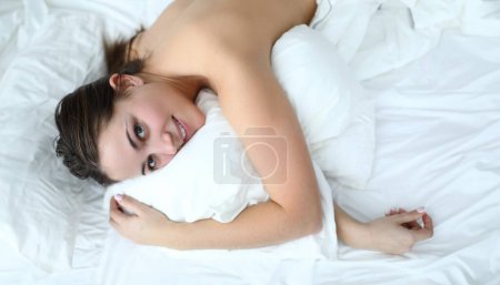 Morgenmädchen entspannt im weißen Bett liegend und lächelnd. Komfortabler Aufenthalt zu Hause Quarantäne. Stimmen Sie sich auf das frühe Erwachen ein. Schlafen hilft, Muskelklemmen zu entschärfen und loszulassen. Hypoallergene Bettwäsche