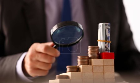 Hand businessman in suit hold lens background looking through money towers closeup search business concept