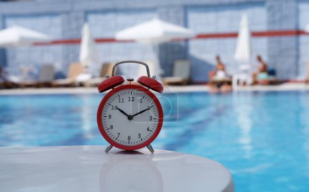 Red analog alarm clock with blurred background of relaxation pool. Resort concept