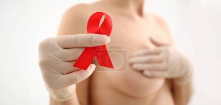 Female hand in gloves holding red ribbon cancer symbol breast closeup on doctor mammology clinic reception. Mammary glands test biopsy implant silicone insert tumor human concept