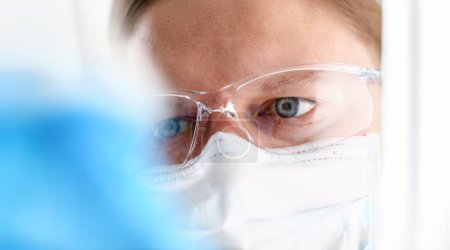 ein Porträt eines jungen Chirurgen Chemiker Arzt blickt auf ein Gefäß mit einer blauen Flüssigkeit und eine Maske wird mit Viren und einem Impfstoff gegen Krankheiten bekämpft.