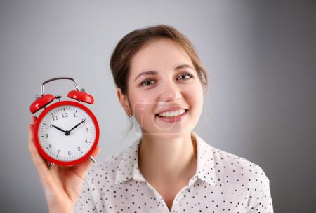 Adult smiling caucasian woman hold red alarm clock on gray background.