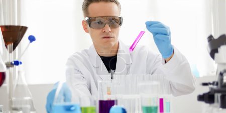 Photo for Male chemist holds test tube of glass in his hand overflows a liquid solution of potassium permanganate conducts an analysis reaction takes various versions of reagents using chemical manufacturing. - Royalty Free Image