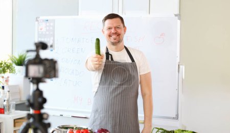 Photo for Smiling Vlogger Chef Recording Green Cucumber. Cook in Apron Cooking Vegetable on Camera. Culinary Recipe Vlog at Kitchen. Organic Raw Vegetarian Food. Man with Ripe Ingredient Looking at Camera Shot - Royalty Free Image