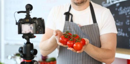 Photo for Vlogger Chef Showing Ripe Tomatoes Ingredient. Man in Apron Recording Big Red Vegetable on Camcorder for Culinary Vlog. Cooking Recipe. Male Hands Holding Fresh and Aromatic Food at Kitchen. - Royalty Free Image