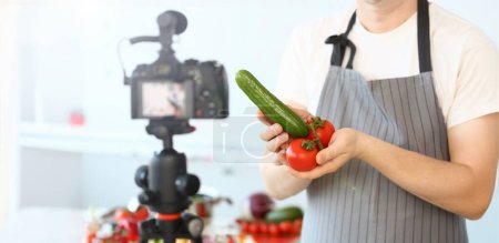 Photo for Comic Culinary Blogger Holding Organic Vegetables. Chef Recording Joke with Cucumber and Tomato for Blog on Camera. Man in Apron Showing Ingredient Assortment for Healthy Food Horizontal Shot. - Royalty Free Image
