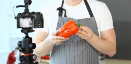 Photo for Chef Vlogger Showing Camcorder Orange Pepper. Man in Apron Recording Ripe Vegetable on Camera for Culinary Vlog in Kitchen at Home. Male Hands Holding Fresh Ingredient Horizontal Photography - Royalty Free Image