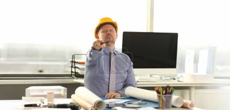 Photo for Serious Architect Working on Construction Outline. Caucasian Engineer Sitting at Office Workplace. Man Wear Formal Shirt, Tie and Yellow Helmet do House Drafting. Businessman Pointing at Camera - Royalty Free Image