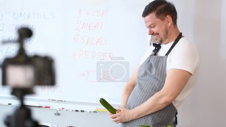 Photo for Smiling Vlogger Chef Recording Cucumber Joke. Cook in Apron Joking with Green Cuke on Camera. Culinary Vlog for Vegan at Home Kitchen. Cheerful Man Vegetable Ingredient for Cooking Vegetarian Food - Royalty Free Image