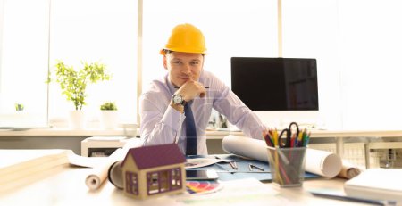 Photo for Young Architect Design Building Plan at Office. Businessman Wearing Shirt, Tie and Yellow Casque Designing House Model on Blueprint Front Shot. Safety Contractor Occupation Concept - Royalty Free Image