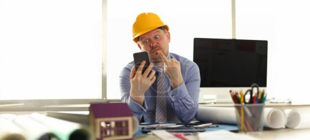 Photo for Architect Express Emotion at Engineering Office. Annoyed Caucasian Constructor in Shirt and Yellow Helm Giving Middle Finger to Mobile Phone in Hand. Angry Building Business Owner - Royalty Free Image