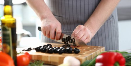 Photo for Professional Chef Cutting Black Olive Ingredient. Man in Apron Chopping by Big Kitchen Knife on Wooden Board. Healthy Culinary Recipe. Cooking Dieting Food at Kitchen Horizontal Photography - Royalty Free Image