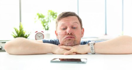 Photo for Man with funny facial expression staring at cellphone lying at table waiting for call concept - Royalty Free Image