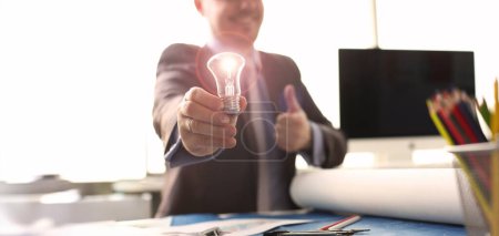 Photo for Inspiration Brand New Idea Originative Symbol. Happy Businessman Holding Traditional Light Bulb. Thumb Up Sign Manager in Suit with Lamp in Hand Sit at Table. Innovation Technology. Accounting Concept - Royalty Free Image