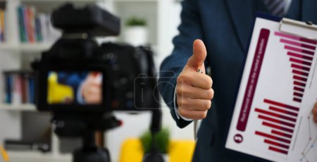 Hombre en traje y corbata mostrar confirmar signo de brazo haciendo videoblog promocional o sesión de fotos en la videocámara de la oficina para trípode primer plano. Vlogger promoción selfie solución o información de gestión asesor financiero
