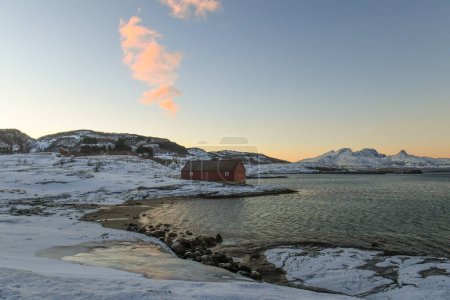 Photo for A traditional norwegian red barn enclosed by rugged mountains and snow-covered beaches in the arctic. Shown during the golden hour of a brief period of daylight near the town of Bodo. - Royalty Free Image