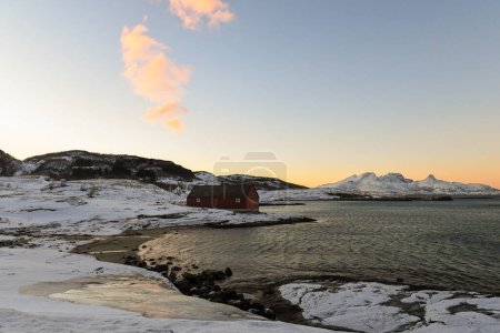 Foto de Un granero tradicional noruego rodeado de montañas escarpadas y playas cubiertas de nieve en el Ártico. Se muestra durante la hora dorada de un breve período de luz del día cerca de la ciudad de Bodo. - Imagen libre de derechos