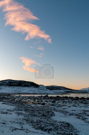 A bright orange glow colours the golden hour sky that serves as a dramatic background for the rugged mountains and snow-covered beaches of arctic norway, near the town of Bodo.