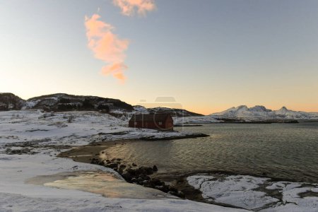 Un granero tradicional noruego rodeado de montañas escarpadas y playas cubiertas de nieve en el Ártico. Se muestra durante la hora dorada de un breve período de luz del día cerca de la ciudad de Bodo.
