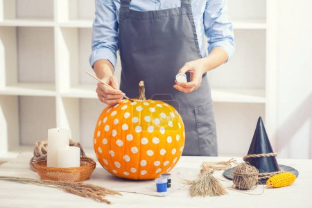Photo for Woman in apron painting Halloween pumpkin, at wooden table, preparing holiday decorations at home - Royalty Free Image