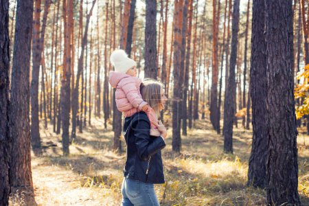 Téléchargez les photos : Mom and little baby daughter are walking in a pine forest. mother carries her baby on her neck - en image libre de droit