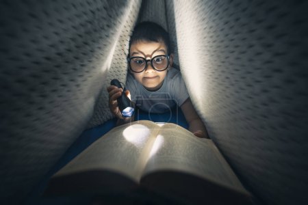 Téléchargez les photos : Petit garçon allongé sur le lit sous la couverture tenant la lampe de poche et le livre de lecture tard dans la nuit - en image libre de droit