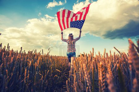Foto de Hermosa joven sosteniendo una bandera americana en el viento en un campo de colza. Paisaje de verano contra el cielo azul. Día de la Independencia 4 de julio - Imagen libre de derechos