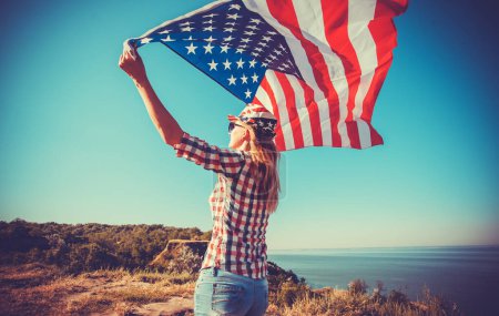 Téléchargez les photos : Belle jeune fille tenant un drapeau américain au vent dans une prairie. Paysage estival contre le ciel bleu. Jour de l'indépendance 4 juillet - en image libre de droit