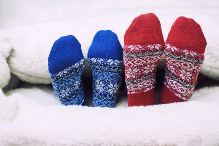 Foto de Christmas red and blue warm socks covered with blanket on white bed. Happy moments of winter holiday. Background, copy space. - Imagen libre de derechos