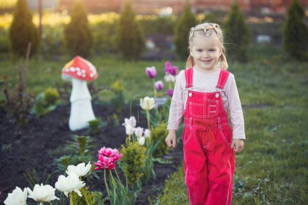 Téléchargez les photos : Une petite fille dans un ensemble rouge près d'un lit fleuri avec des tulipes - en image libre de droit
